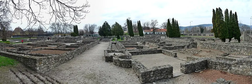 A panorama across the ruin field under a grey sky, with knee-high walls of rough stone enclosing rooms of bare earth, a gravel path running between them and tall cypresses behind.