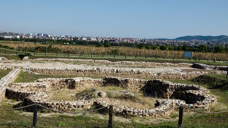 Low stone foundations of a building with rounded rooms, behind a rope fence on wooden posts, with an information board to the right and the tower blocks of a modern town on the horizon.
