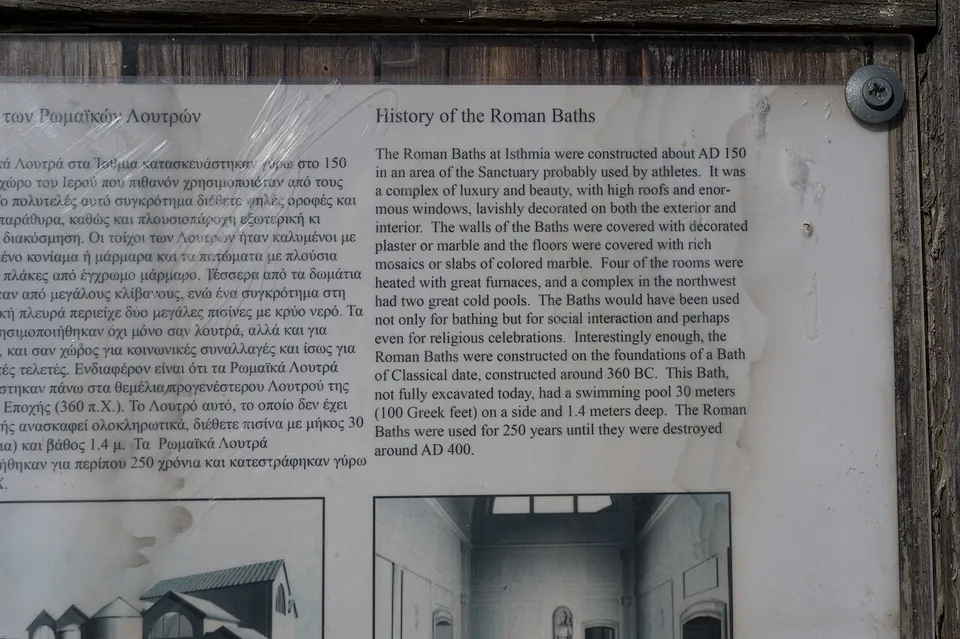 An information board behind scratched plastic with Greek and English text headed "History of the Roman Baths", and drawings of a reconstructed bath building below.