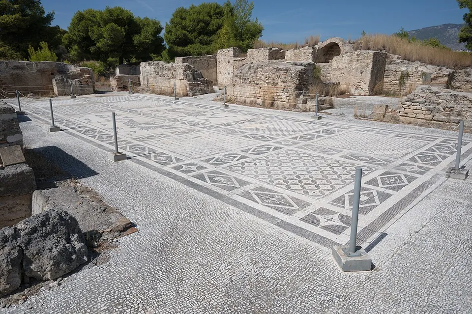 A large floor of black-and-white mosaic in geometric patterns, fenced by low posts, with ruined walls of squared stone and an arch behind it and pine trees beyond.