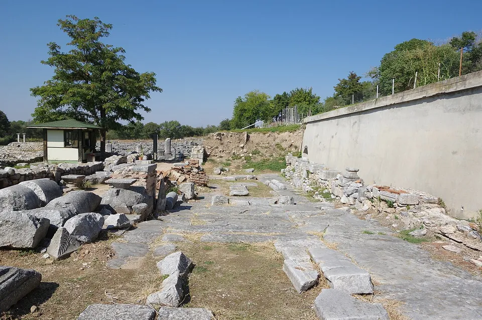 A broad road of large worn paving stones running between heaps of fallen blocks and a modern retaining wall, towards a small kiosk and trees.