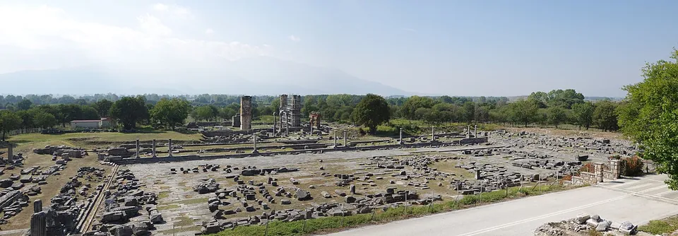 A panorama over a broad excavated square of paving, foundations and scattered blocks among trees, with tall brick piers of a ruined building standing in the distance and mountains beyond.
