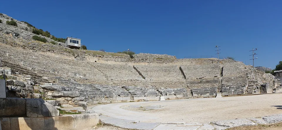 A large theatre of stone seating rows set into a rocky hillside, around a flat sandy orchestra, under a deep blue sky.