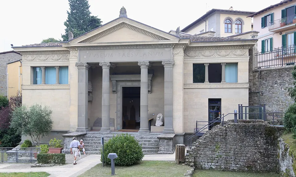 A pale building in the form of a classical temple, with a pediment and columns framing its entrance, a Latin inscription along the frieze and a short flight of steps up from a paved path.