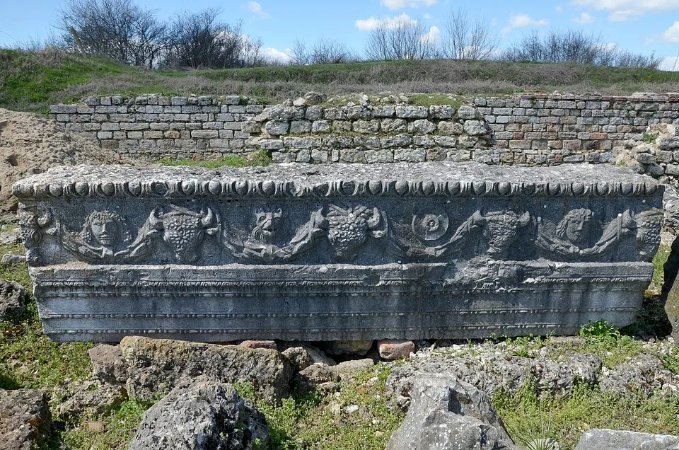 A long carved stone beam lying on the ground, its face decorated with garlands, faces and bulls' heads under a band of moulding, with a wall of squared stones behind.