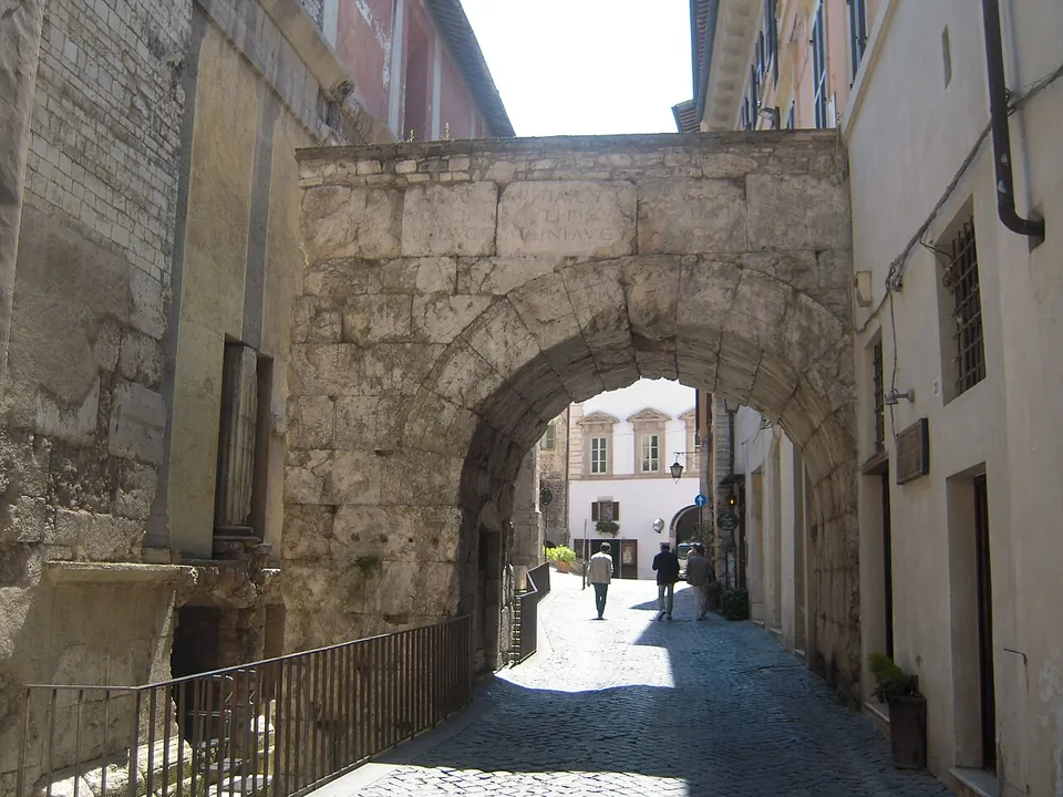 A single round arch of large rough limestone blocks spanning a narrow cobbled street between tall houses, with a railing along a sunken strip on the left and people walking beyond.
