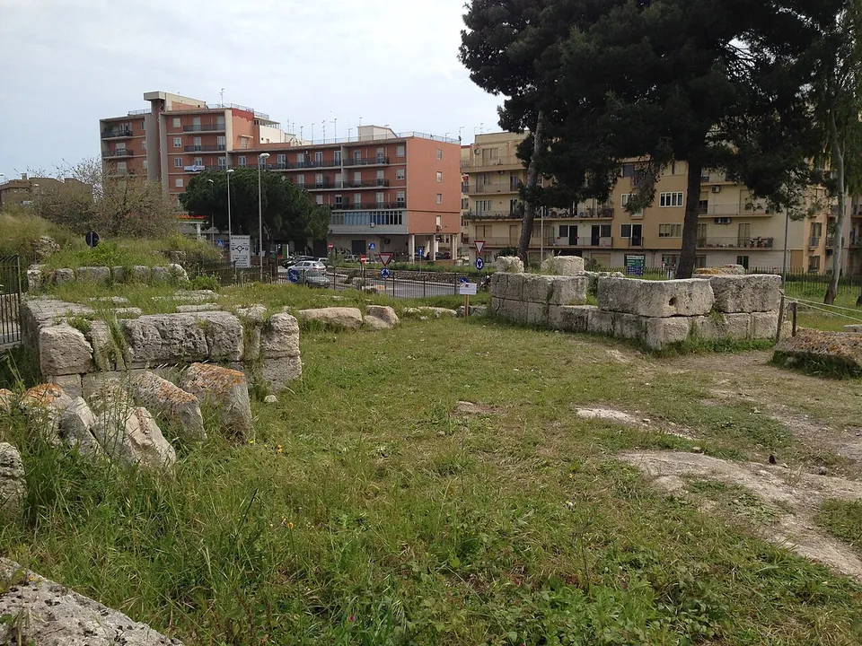 Two low clusters of large, roughly dressed limestone blocks in a grassy strip, a few metres apart, with apartment blocks and trees behind.