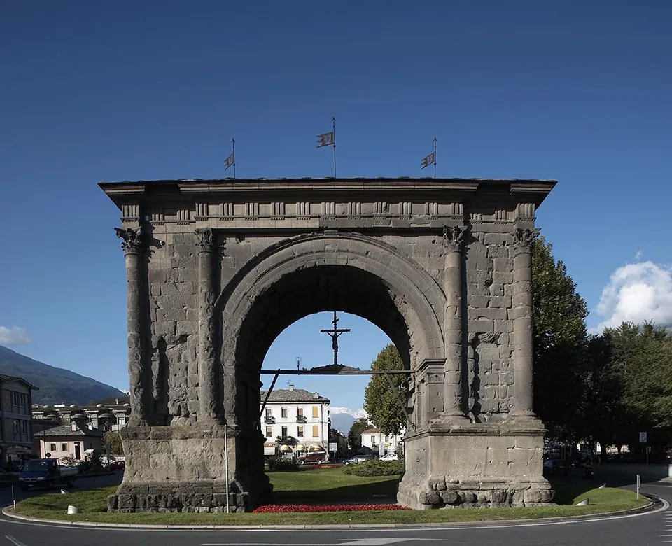 A free-standing arch of dark grey stone with a single high opening, engaged columns with carved capitals on its front and a pitched roof in place of an attic, standing on a lawn in the middle of a roundabout, a crucifix hanging inside the arch.