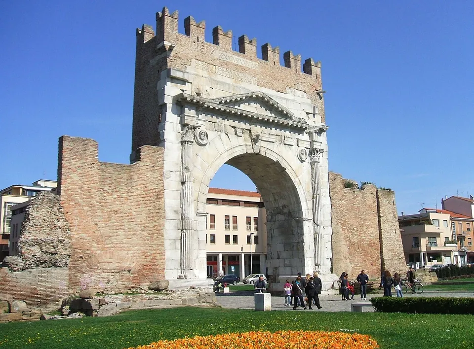 A tall single-arched gate faced in pale stone, with columns either side of the opening and a pediment above it, topped by a band of brick with swallowtail battlements; low stretches of brick wall run off on both sides, with a lawn and a flowerbed in front.