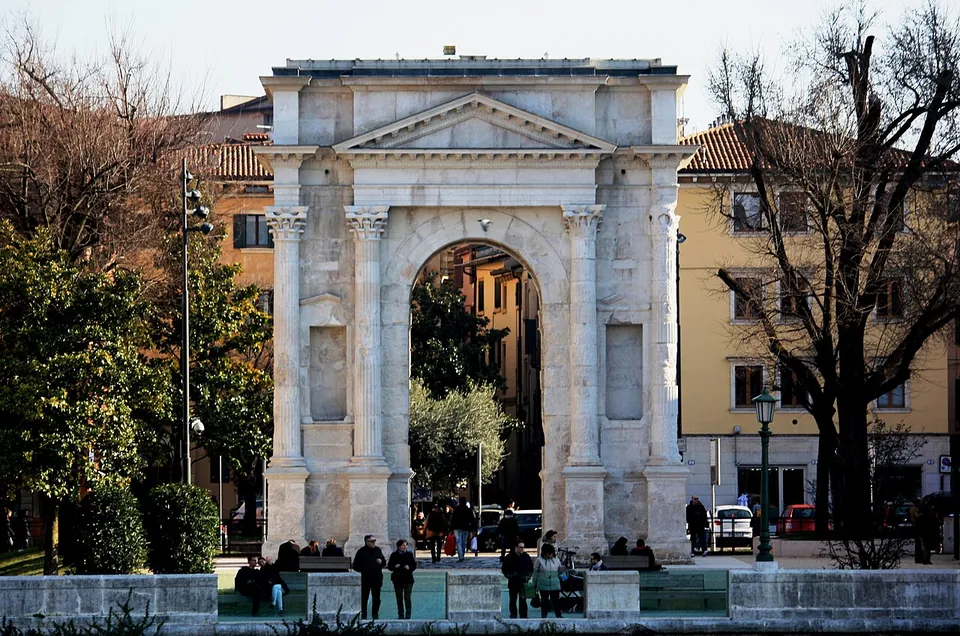 The arch seen from across the river behind a low wall and shrubs, its pale stone face with columns, niches and pediment set between bare trees, with houses and a few people in the square behind it.