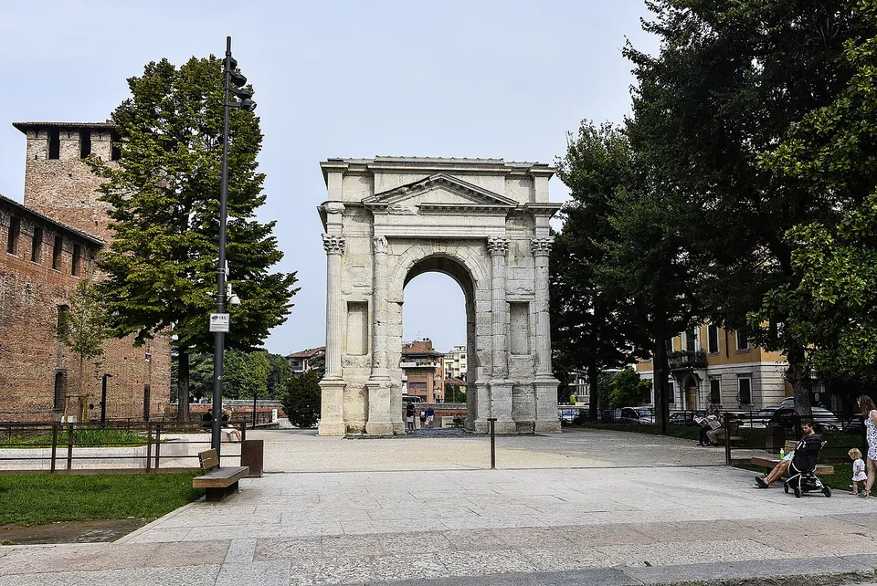 A wide paved square with lawns, benches and a street lamp, a single pale stone arch standing in it between tall trees, and a crenellated brick tower of a medieval castle at the left.