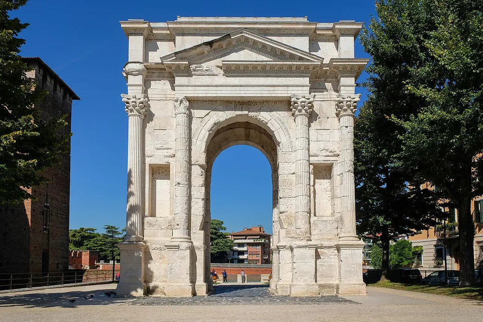 A free-standing arch of pale stone on a paved square under a blue sky, its front framed by four fluted columns with ornate capitals, a pediment over the central opening and a tall narrow niche on each side; trees and a brick castle wall stand beside it.