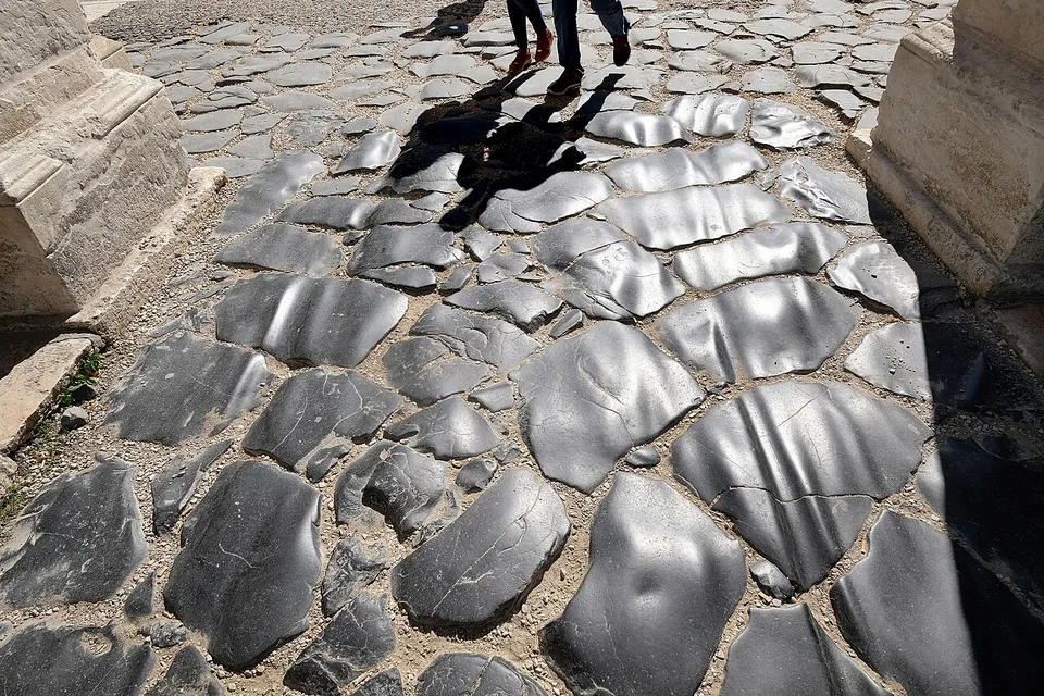 Close view of a road surface of large, irregular, dark polished paving stones between the stone bases of the arch's piers, with the shadows of two walkers falling across it.