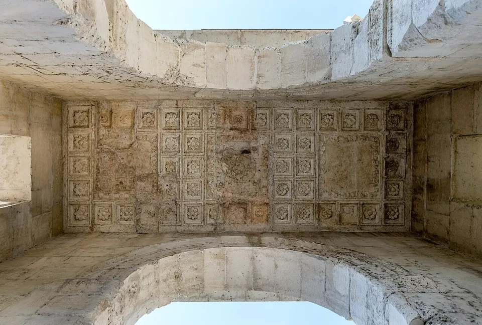 Looking straight up inside the arch at a flat stone ceiling divided into rows of small square coffers, its surface worn and uneven, framed by the curves of the two main arches against the sky.