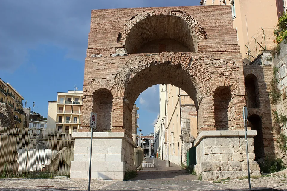 A tall arch of red-brown brick spanning a narrow street between modern buildings, with a smaller arched niche on each side of the opening and a second, recessed arch above it, the whole standing on two bases of pale squared stone blocks; a railing fences a paved area to the left.