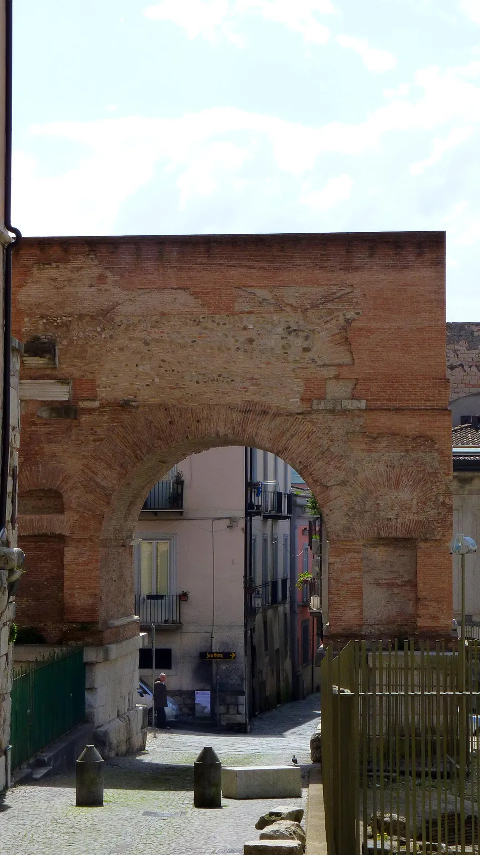 The arch seen head-on down a short paved lane, its brick face weathered and patched, with houses visible through the opening and a wooden fence and green railing in the foreground.