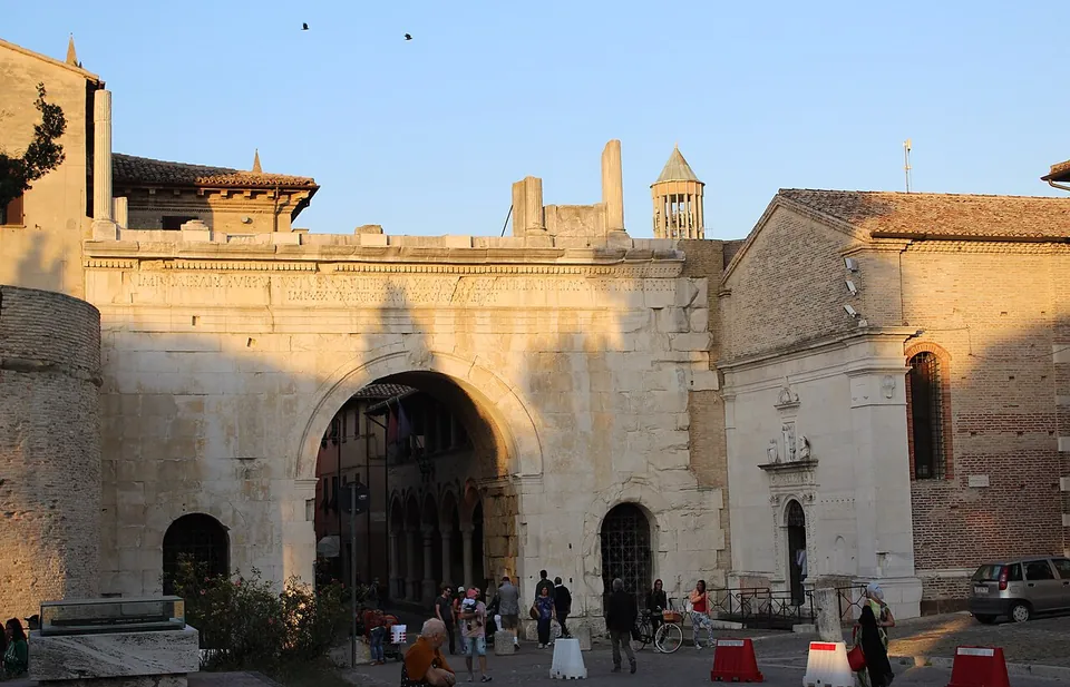 A broad gate of pale squared stone with one large round-headed central arch and two small arched side openings, ruined stumps of masonry along its top and a bell tower rising behind, a church front with a carved doorway built against its right side, and people and bicycles in the square in front.
