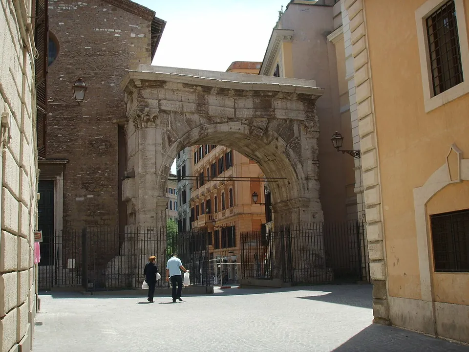 A single round arch of pale weathered stone with flat pilasters and a plain band across the top, standing in a narrow paved street between a brick church wall and a pale apartment building, with iron railings under the opening.