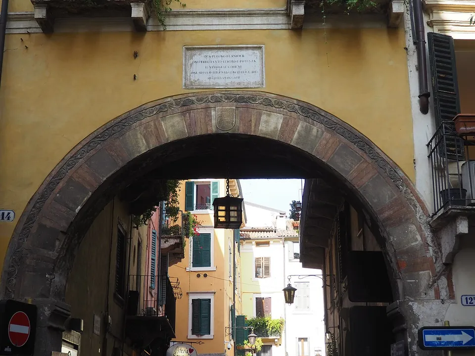 A wide round arch with a band of carved stone ornament in an ochre-painted building, a marble plaque above it, opening on a narrow lane with shuttered windows and a no-entry sign.
