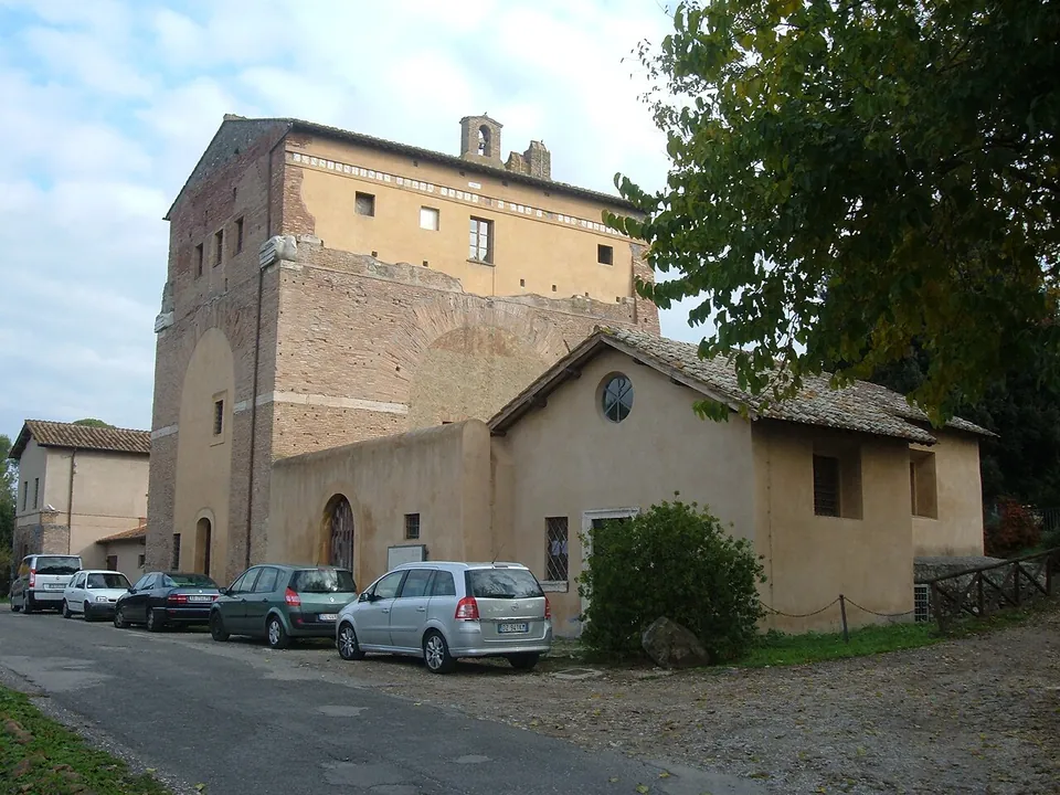 The tall brick block of the farmhouse seen from the lane, with a lower ochre-plastered house built against it, cars parked along the front and trees to the right.
