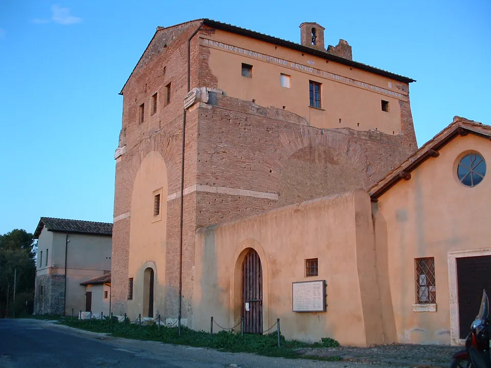 A corner of the building in evening light, its red-brick upper walls rising above a plastered lower range with an arched, barred doorway and a round window.