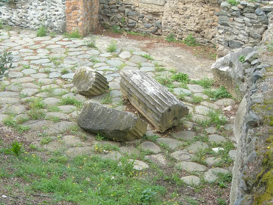 Paving of irregular grey stone blocks with grass growing between them, three fallen fragments of fluted stone column lying on it, and low rubble walls behind.