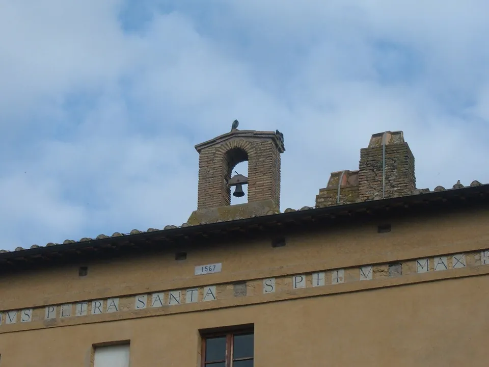 The top of the ochre wall under the eaves, with a small brick bell-gable and chimneys on the roof, a plaque reading 1567 and a band of lettering in which "PETRA SANTA" can be read.