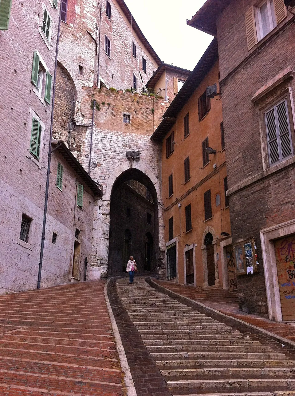A long flight of brick-paved steps climbing between tall old houses to a high, pointed archway in a wall of pale stone.