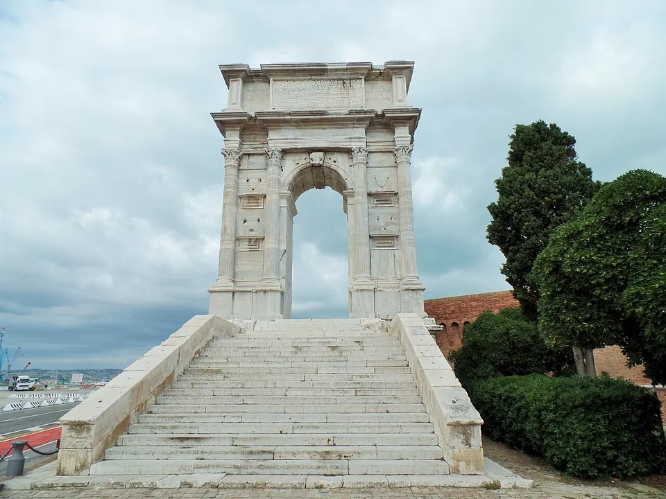 A tall, narrow single arch of pale marble on a high podium, framed by pairs of fluted columns and topped by a plain attic, reached by a broad flight of stone steps, with trees to one side and the port behind.