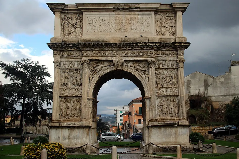 A tall single-arched gateway of pale marble, its whole face covered in carved relief panels between engaged columns, with an inscription panel at the top, standing in a small fenced lawn with houses and a cloudy sky beyond.