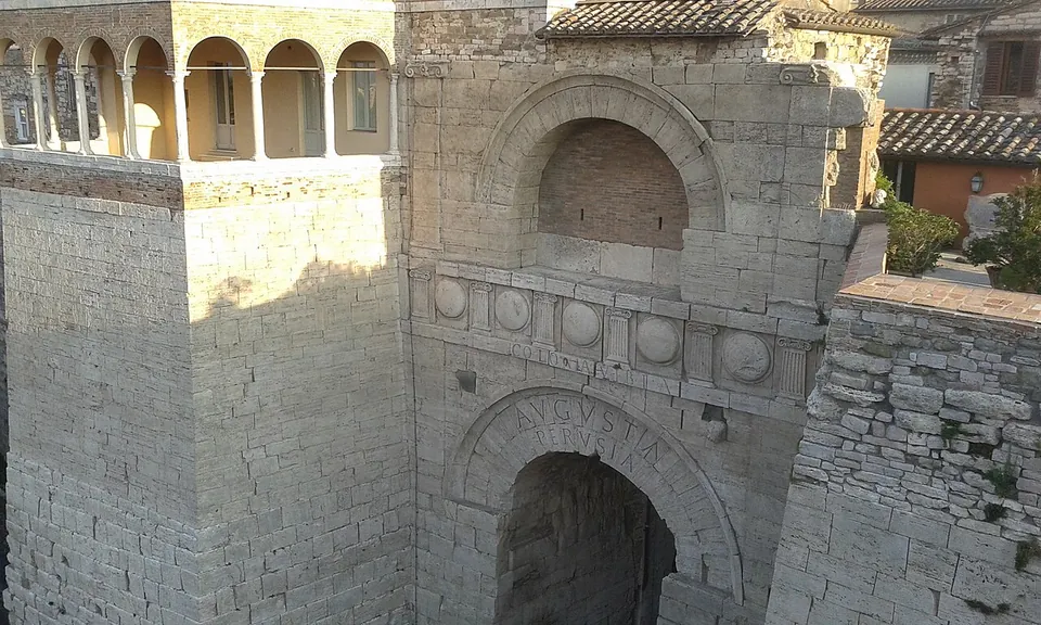 The central front of the gate seen from a window opposite, in evening light. Large letters reading AVGVSTA PERVSIA are cut into the stones of the arch; above it runs a band of round shields between small pilasters, with the word COLONIA cut along its lower edge, and higher still a smaller blind arch. The arcaded loggia on the left-hand tower is at the top left.