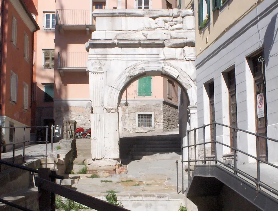 A single round arch of pale stone with a plain block of masonry above it, wedged between old town houses at the top of a short flight of steps with metal railings.