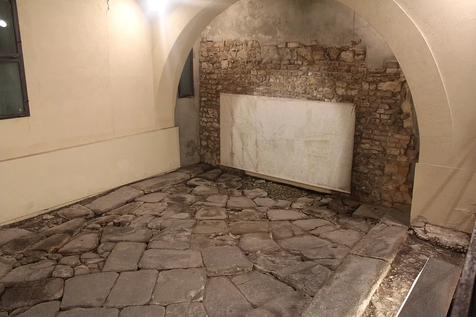 A floor of large, irregular grey paving stones fitted closely together, edged on one side by a row of long kerb slabs, in a white-walled vaulted room with a wall of rough stone and brick at the back and a pale panel leaning against it.