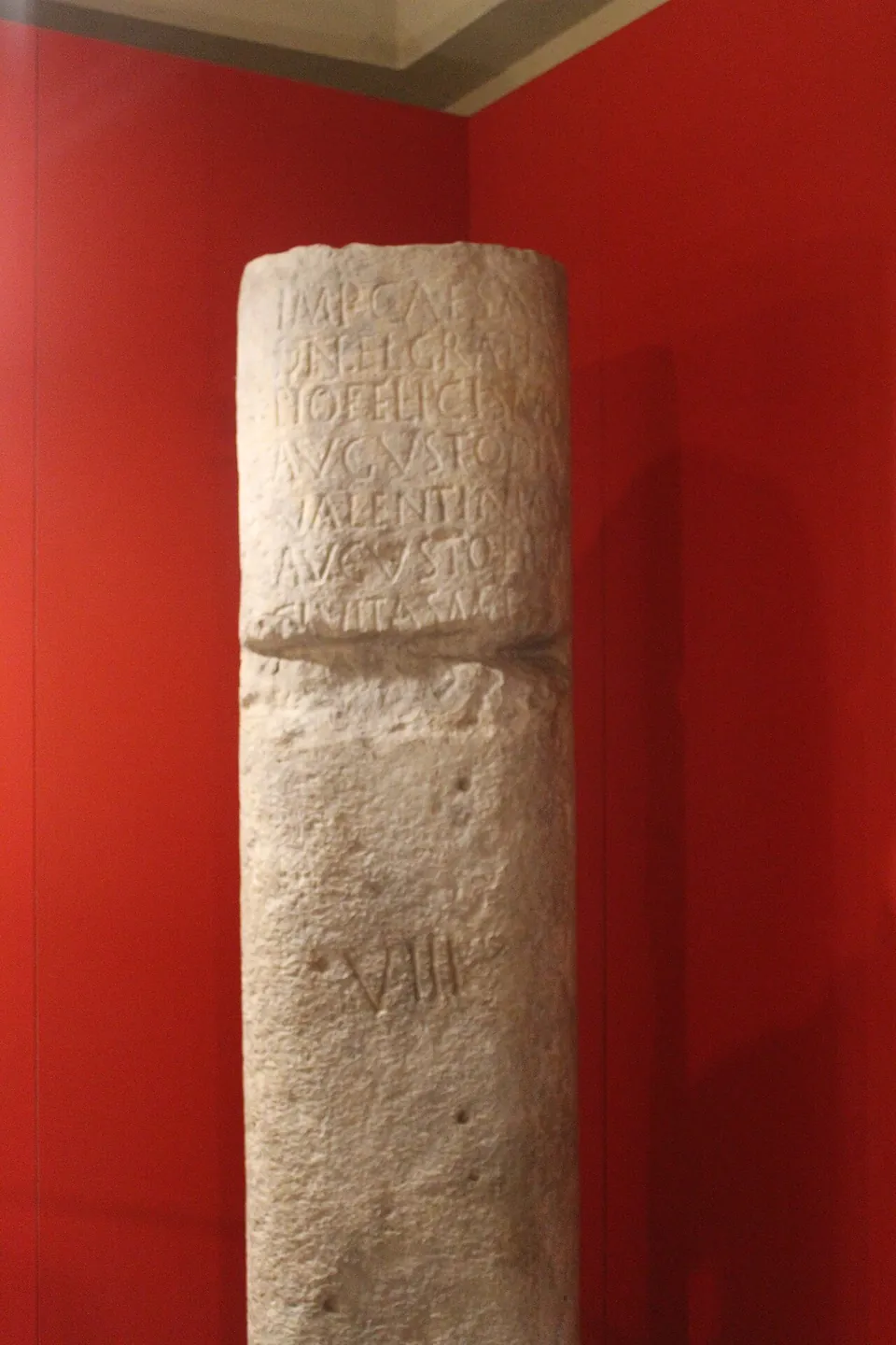 A tall stone column, broken across near the top, standing against a red wall; worn lines of carved Latin letters run round its upper part and the numeral VIII is cut lower down.