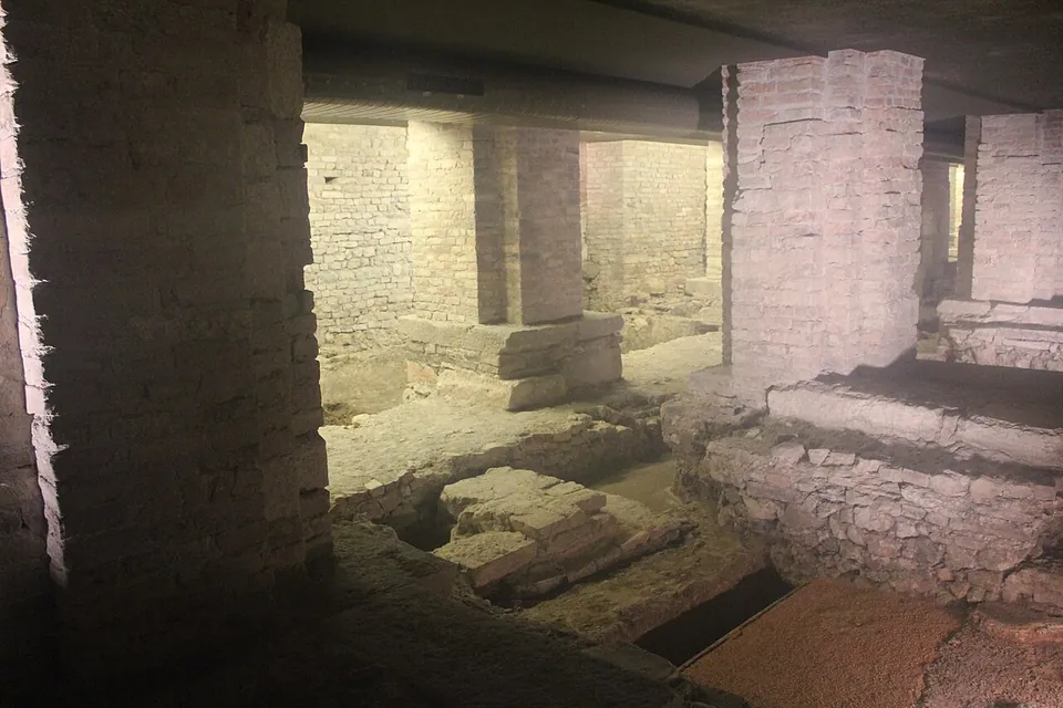 Dimly lit low walls of rough stone and the edges of old floors among modern brick piers, with a dark passage leading away under a concrete ceiling.
