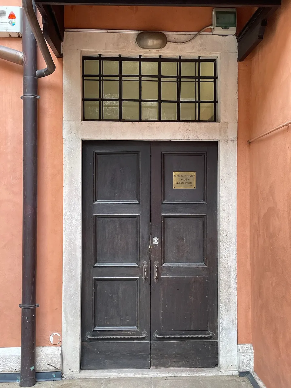A dark wooden double door with a small brass plate, set in a stone frame under a barred glass transom in an orange-plastered wall.