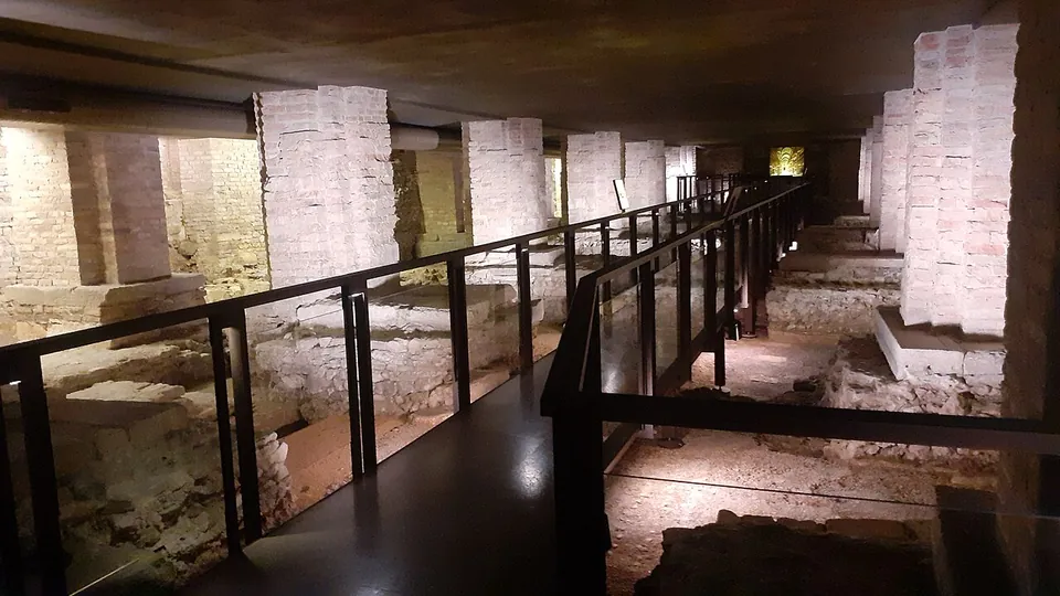 A long, low underground hall beneath the cathedral, where a black-railed walkway runs between square brick piers rising to a concrete ceiling, with excavated stone wall footings and patches of floor lying below the walkway on both sides.