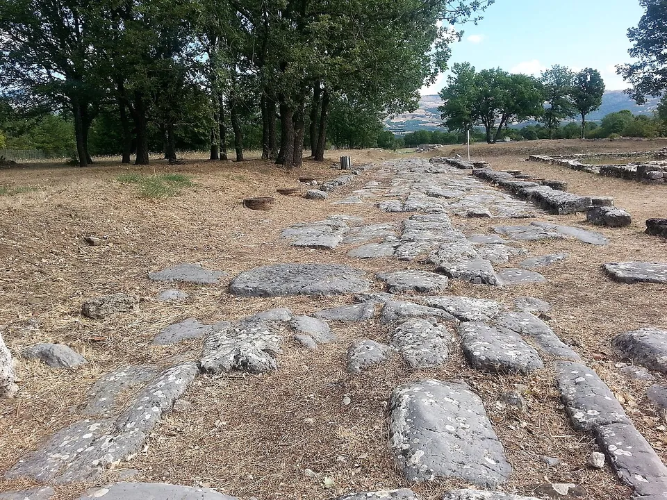A street of large irregular paving stones running straight through dry grass under a line of oak trees, with low kerb walls along its edge.