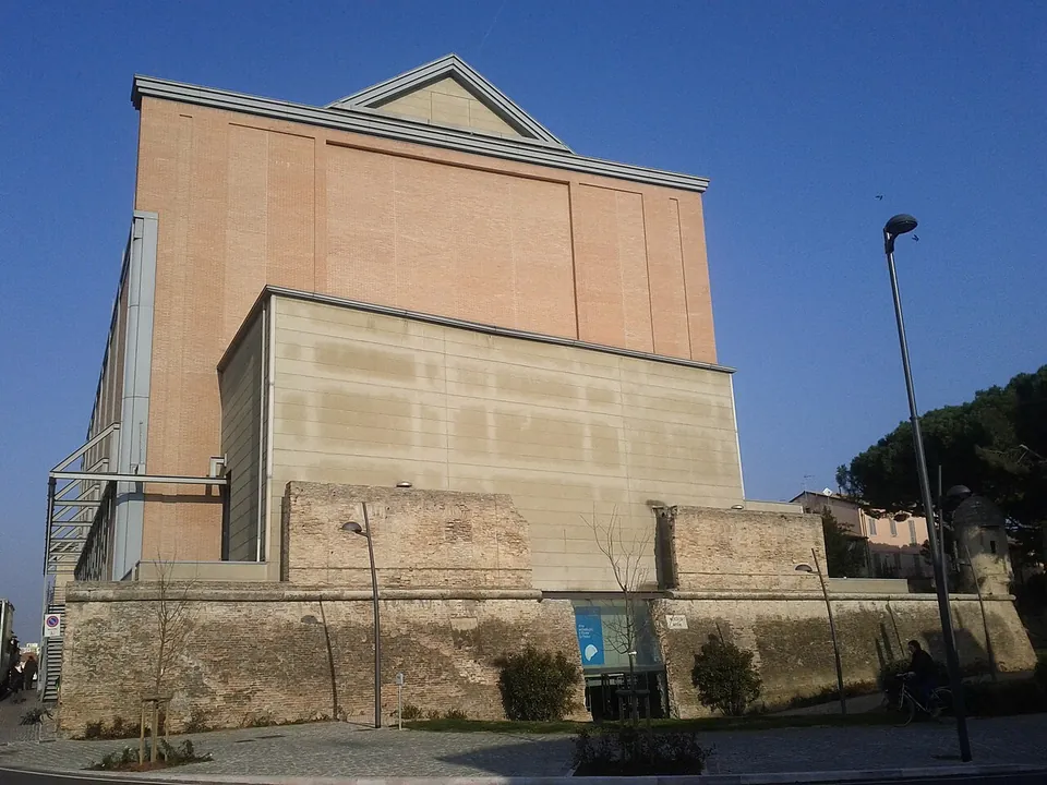The tall, plain block of the La Fenice theatre rising above an old brick-and-stone wall, with a glazed doorway and a blue banner at the foot of the wall.