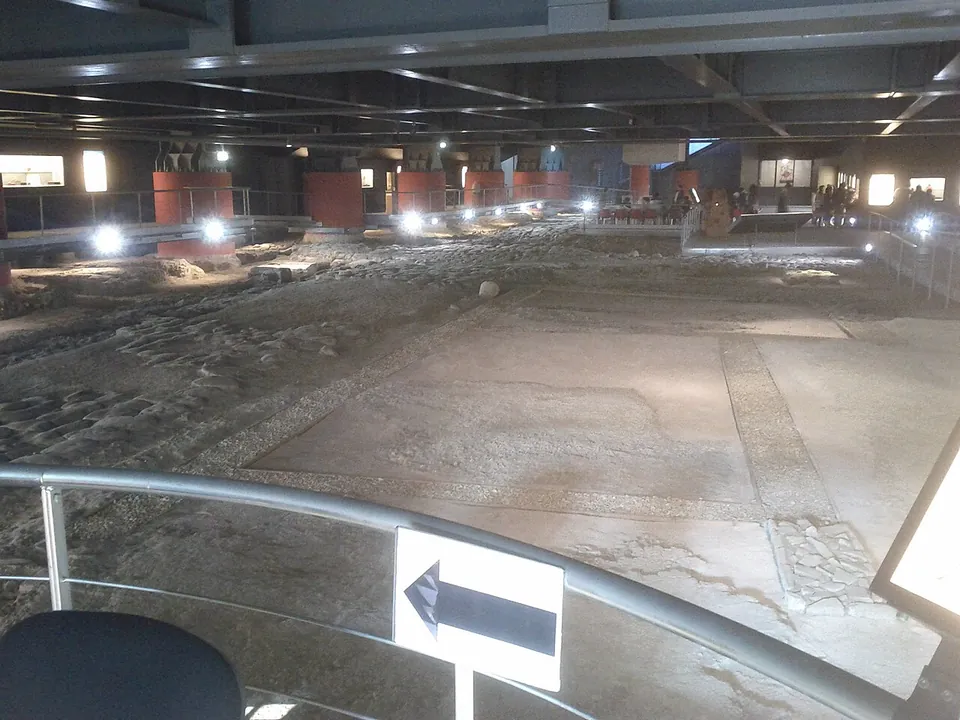 A long, low underground hall under a steel ceiling, seen over a railing from the entrance walkway. Smooth pale floor surfaces divided by narrow borders fill the foreground, a band of rough stone paving runs along the left, and walkways lit by spotlights edge the hall past red-painted pillars and lit display cases.