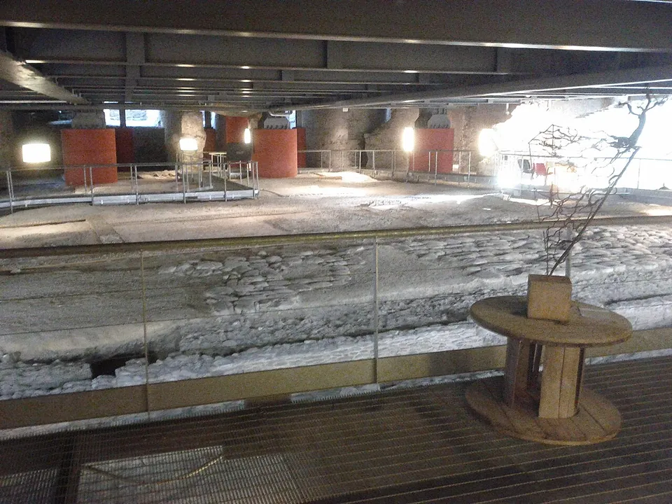A broad band of stone paving crossing the underground hall behind a railing, seen from a metal-grid walkway, with red pillars and spotlights in the background.