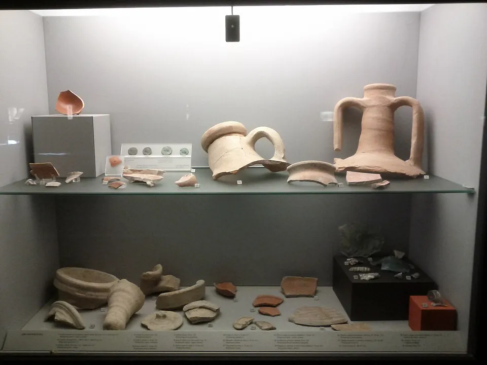 A lit glass display case with two shelves of pottery, among them a large two-handled jar, the neck of another jar and many broken pieces, each with a small label.
