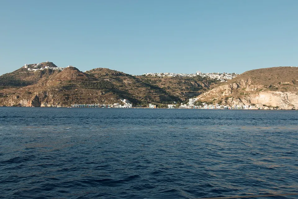 Brown hills rising from a calm blue sea, with a white village on the left-hand summit, another white village along the ridge on the right and a row of small houses at the water's edge below.