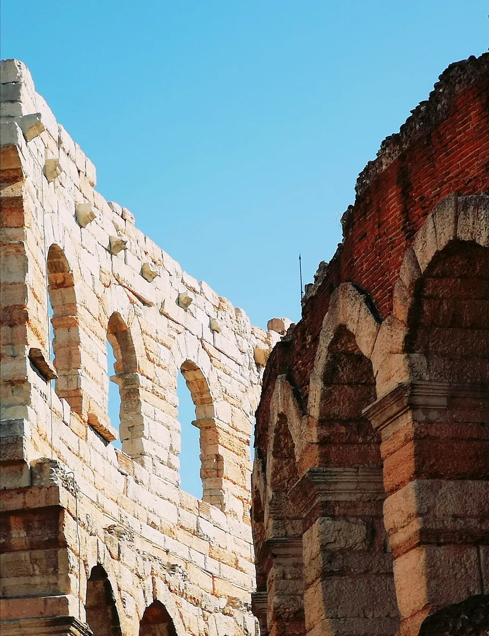 Close view of a high wall of large pale stone blocks with arched openings and rows of projecting stone brackets, beside the top of an arcade of stone arches set in red brick.
