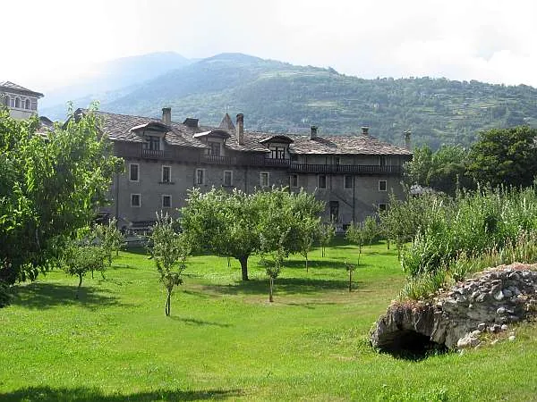 A lawn planted with young fruit trees, with a long building with a stone-slab roof behind it and a low stretch of rough masonry in the grass on the right.