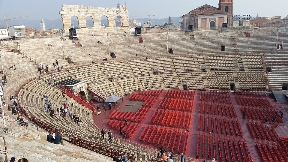 Inside the amphitheatre seen from high on the stone seating, rows of red seats set out in blocks on the oval floor, a covered stage area at one end, the stone tiers sweeping round, and the arched fragment of the outer wall standing above the far side.
