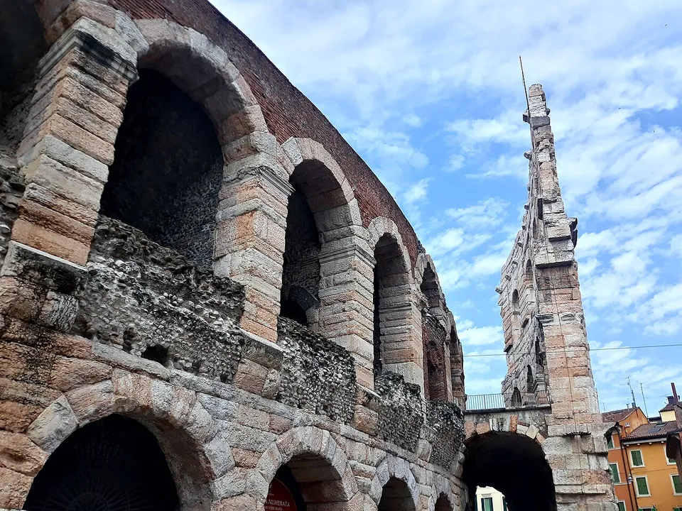 Looking up along the curving arcades of the amphitheatre, two storeys of round arches in pale and pinkish stone blocks, with a much taller, narrow fragment of wall rising beyond them against a blue sky.
