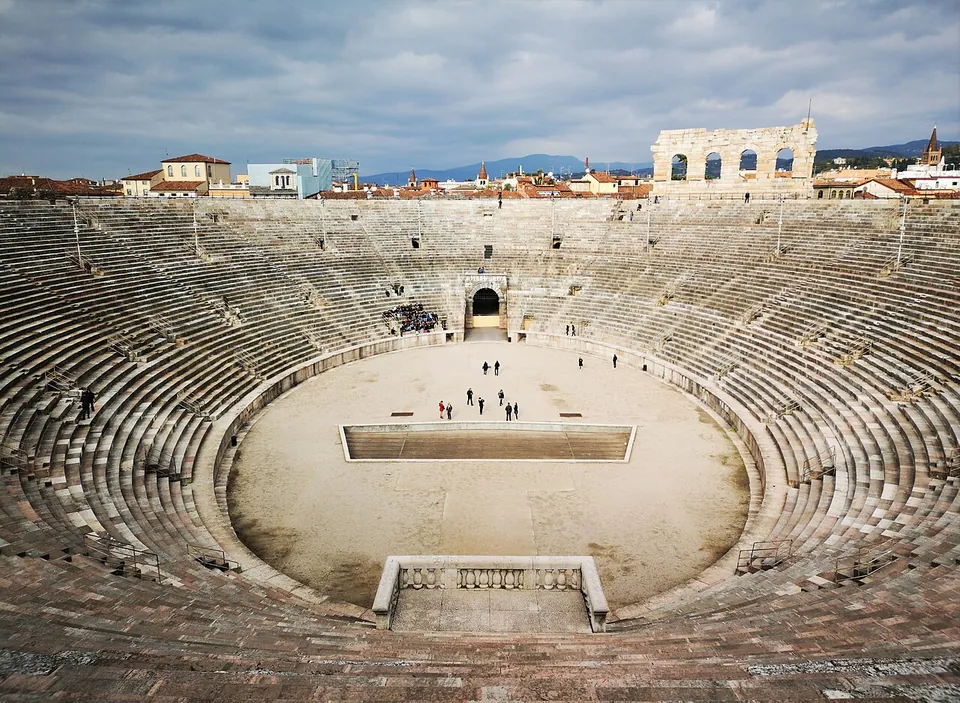 Inside the amphitheatre, rows of stone steps sweep round an oval sand-coloured floor where a few people stand; beyond the top of the seating a fragment of a taller wall with arched openings stands against the sky above the rooftops.