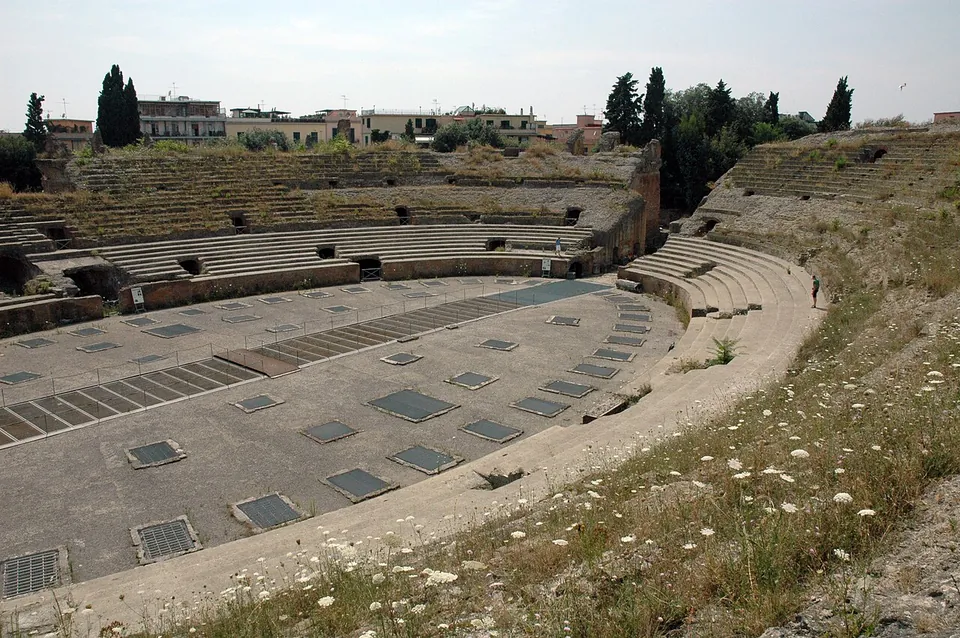 The oval floor of the arena, set with rows of rectangular openings covered by grilles, ringed by a low wall and by banks of stone seating overgrown with grass, with cypresses and modern buildings beyond.