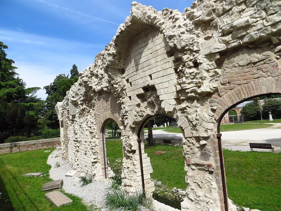 A ruined wall of rough rubble and dressed stone with brick-edged arched openings, some of them barred, standing on a lawn beside a gravel path in a park.